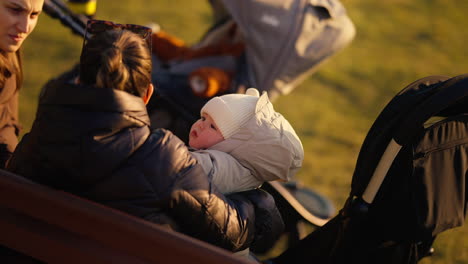 Portrait-of-a-baby-in-its-mother's-arms-on-a-bench-in-a-public-city-park,-on-a-spring-evening