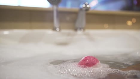 Pink-bath-bomb-dissolves-in-foamy-bubbly-water-in-tub,-close-view