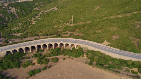 Aerial-view-following-car-traffic-moving-over-bridge-during-daytime,-an-stone-arch-bridge-in-the-mountains-north-of-the-coastal-town-of-Tarifa-in-Andalusia,-Spain
