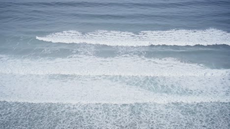 Foamy-ocean-waves-roll-towards-coastline,-view-from-above