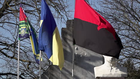 The-regional-flag-of-Kherson,-the-national-Ukraine-flag-and-the-red-and-black-flag-of-Ukrainian-resistance-fly-next-to-a-statue-of-Taras-Hryhorovych-Shevchenko-in-slow-motion