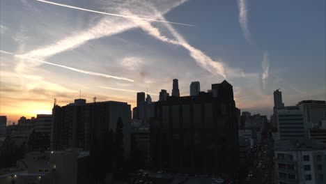 Eine-Aufsteigende-Luftaufnahme-Zeigt-Die-Silhouette-Der-Innenstadt-Von-Los-Angeles-Mit-Einem-Epischen-Orange-goldenen-Sonnenuntergangshimmel-Mit-Kondensstreifen