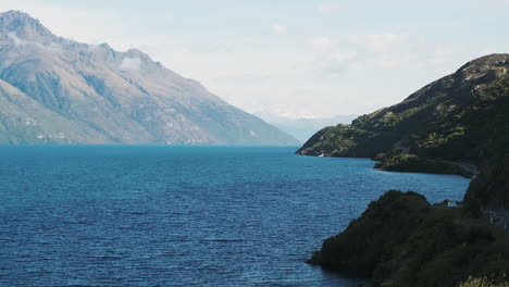 Car-driving-on-winding-mountainous-New-Zealand-road-alongside-bright-blue-lake