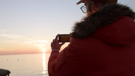 Man-in-an-orange-jacket-taking-pictures-with-his-smartphone-of-the-sun-setting-behind-the-ocean