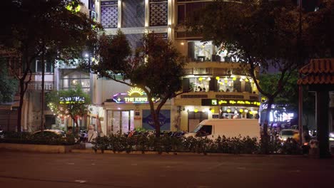 Verkehrsaktivität-Mit-Ladengeschäften-Im-Hintergrund,-Aufgenommen-Aus-Einem-Park-Bei-Nacht-In-Ho-Chi-Minh-Stadt,-Vietnam