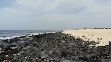 Aerial-Low-Flying-Over-Dark-Rocks-Creating-Breakwater-Beside-Sandy-Beach-At-Cape-Verde