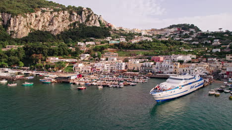 A-wide-drone-shot-circling-a-large-ferry-in-Marina-Grande-on-the-island-of-Capri-in-Campania,-Italy,-with-boats-and-tourists-in-the-port-and-a-lush-island-backdrop,-rocky-cliffs-and-pastel-buildings