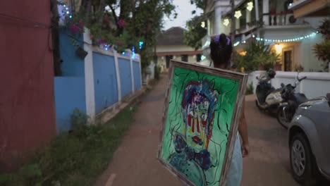 A-shot-following-an-Asian-Female-walking-down-a-residential-road-in-the-early-evening,-the-Artist-holding-a-picture-frame-with-her-most-recent-abstract-painting-of-a-face,-Goa,-India