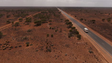 Aerial-view-of-camper-van-on-empty-road-in-Australia-surrounded-by-red-desert-during-cloudy-day