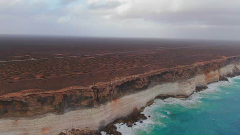 Cinematic-drone-shot-of-Campervan-on-empty-road-in-Nullarbor-SA,-Australia-during-cloudy-day---Ocean-waves-crashing-against-steep-rocky-coastline