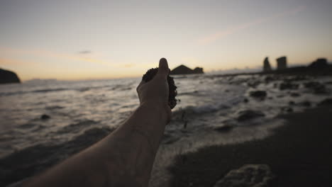 Male-hand-picks-up-sand-on-beach-by-rocky-Azores-coast-at-dusk,-POV