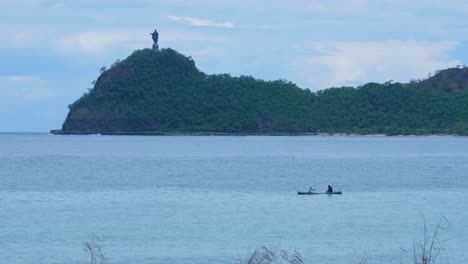 Pescador-Timorense-Local-En-Una-Canoa-De-Pesca-Tradicional-Con-Estatua-De-Cristo-Rei-En-La-Distancia-En-La-Ciudad-Capital-Dili,-Timor-Leste,-Sudeste-De-Asia
