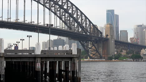 Ein-Mann-Geht-über-Einen-Bretterweg-Am-Hafen-Entlang,-Im-Hintergrund-Die-Stadt-Sydney