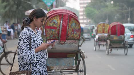 Una-Mujer-Usa-Un-Teléfono-Al-Lado-De-La-Carretera-Mientras-El-Tráfico,-Incluidos-Los-Rickshaws,-Pasa-Junto-A-Ella-En-Una-Carretera-Muy-Transitada-En-Dhaka,-Bangladesh