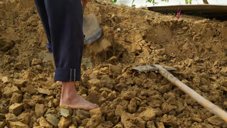 A-barefoot-man-uses-a-hoe-to-loosen-new-clay-soil,-a-raw-material-for-traditional-pottery,-under-the-sun,-depicting-hard-labor-in-rural-life