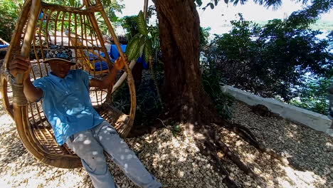 Young-man-sitting-with-blue-tshirt-and-cap-on-his-head,-has-fun-on-a-wooden-swing