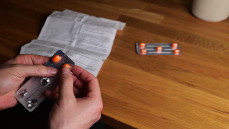 Hands-holding-blister-pack-of-orange-medication-tablets-with-person-reading-medicine-guide-sheet-at-wooden-table