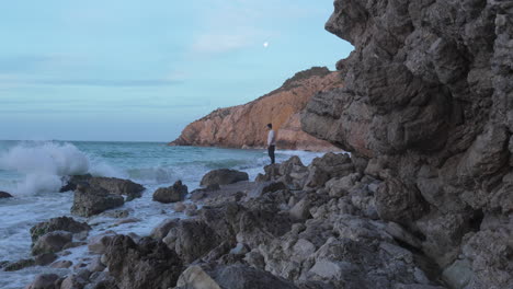 Man-walks-around-cliffs-next-to-blue-moon-sea,-wild-waves-crashing-as-photo-is-taken