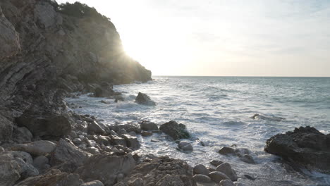 camera-pan-right,-rocky-sea-coast-during-early-morning-as-waves-come-onshore-and-sun-flare-hits-lens