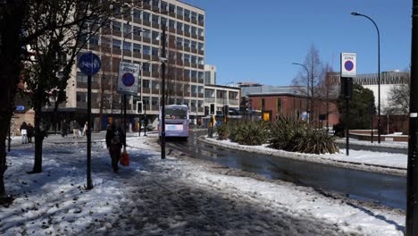 Bus-driving-through-snowy,-sunny-street-and-woman-walking,-Sheffield