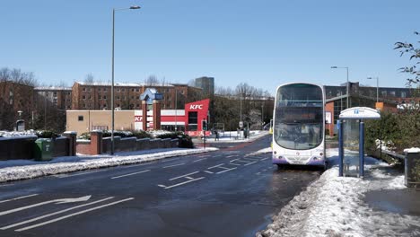 Bus-leaving-bus-stop-and-traffic-driving-past-on-sunny,-snowy-road,-Sheffield