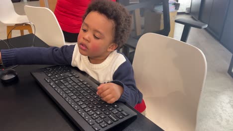 Expressive-three-year-old-black-child-mimics-mother-at-office-desk,-typing-on-computer-keyboard