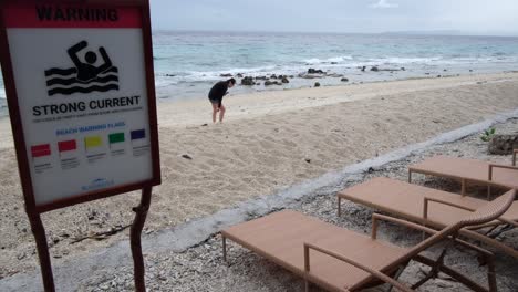 Primer-Plano-De-Una-Fuerte-Señal-De-Advertencia-De-Playa-Actual-Y-Tumbonas-Vacías-En-La-Playa-Con-Un-Turista-Solitario-En-Segundo-Plano-En-Cebu,-Filipinas