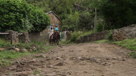Afrikaner-Traben-Mit-Pferden-Auf-Felsigem-Weg-Am-Steinhaus-Im-Dorf-Lesotho-Vorbei