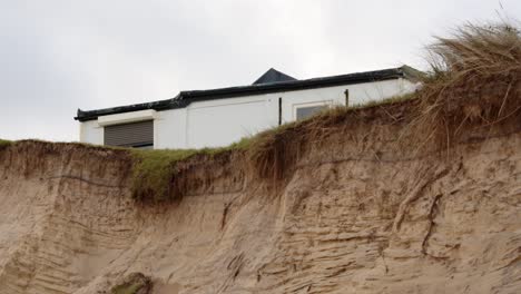 Erosión-Costera-De-Sandunes-En-La-Playa-De-Hemsby-Con-Casa-Al-Borde-De-Un-Acantilado,-Plano-Medio