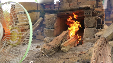 Low-wide-view-of-blazing-wood-fire-in-kiln,-fan-blows-fire-and-smoke,-Than-Ha-Vietnam-Hoi-An