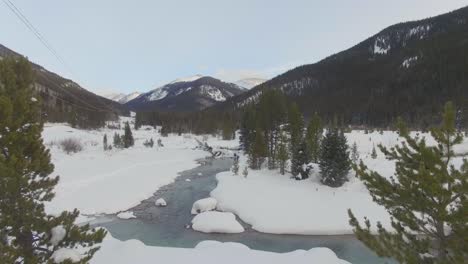Slow-fly-through-small-trees-over-winding-river-in-snow-covered-mountain-valley