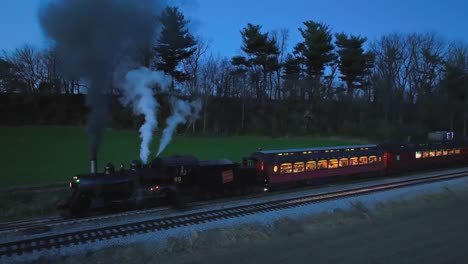 A-Drone-Parallel-Night-View-of-a-Steam-Passenger-Train-Stopped-in-Farmlands-Blowing-Lots-of-Smoke-Seeing-the-Lights-in-the-Coaches