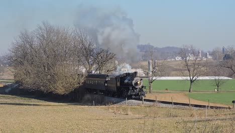 A-Restored-Steam-Passenger-Train-Passes-By-Blowing-White-and-Gray-Smoke,-Traveling-Thru-Rural-Countryside-on-a-Sunny-Winter-Day