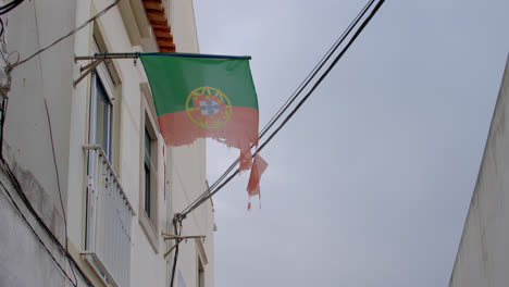 Tattered-flag-of-Portugal-blowing-in-the-wind-in-an-alleyway-in-Nazare,-Portugal
