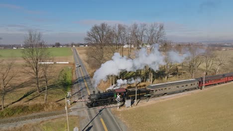 An-Aerial-Side-View-in-Slow-Motion,-of-a-Steam-Passenger-Train-Approaching,-Blowing-Smoke,-while-Traveling-Thru-the-Countryside,-on-a-Sunny-Winter-Day,-on-a-Sunny-Winter-Day