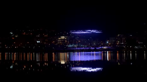 Drones-Elevándose-Hacia-El-Cielo-Nocturno-Sobre-El-Paisaje-Urbano-Reflejado-En-El-Lago-De-Agua-Para-Un-Hermoso-Espectáculo