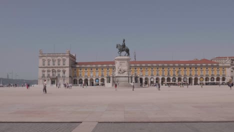 Wide-angle-shot-of-tourists-and-locals-walking-past-statue-and-historic-buildings-in-Lisbon-famous-Praça-do-Comércio,-Portugal-Europe