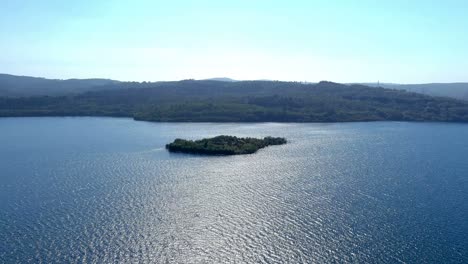 Pequeña-Isla-Con-árboles-Verdes-En-Medio-Del-Lago-Con-Bosques-De-Pinos-En-El-Fondo-Y-El-Cielo-Azul-Sin-Nubes-En-Un-Día-Soleado