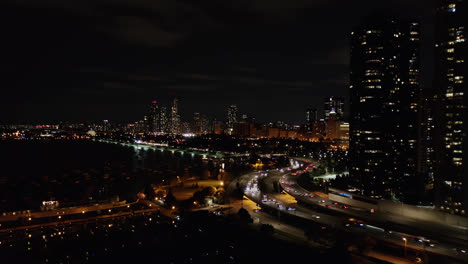 Aerial-drone-shot-of-traffic-movement-over-a-bridge-along-the-seaside-with-the-view-of-high-rise-office-and-residential-buildings-in-the-background-in-Chicago,-USA-at-night-time