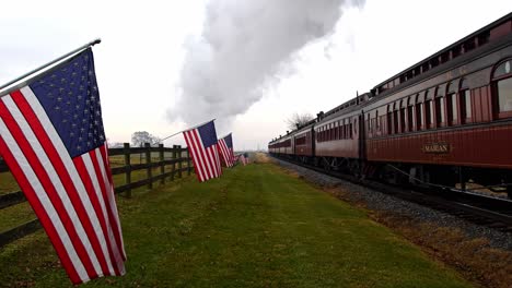 A-View-of-a-Line-of-Gently-Waving-American-Flags-on-a-Fence-by-Farmlands-as-a-Steam-Passenger-Train,-Blowing-Smoke-and-Steam,-Approaches-on-a-Winter-Day