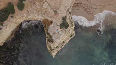 Young-adult-drone-operator-lying-on-top-of-Benagil-rocky-coastal-cliff-with-Atlantic-ocean-below,-Rising-aerial-view