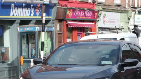 Cork-city-street-on-a-sunny-day,-storefronts,-Domino's-pizza-and-phone-booth-with-slow-moving-traffic