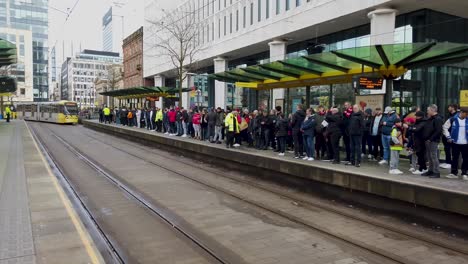 Football-fans-waiting-for-a-tram-on-a-crowded-platform-in-Manchester-City-Centre-and-on-their-way-to-a-football-match-at-Old-Trafford-Stadium,-Manchester,-UK