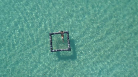 Girl-relaxing-in-an-inflatable-pool-in-the-ocean,-Koh-Kood,-Thailand-DRONE-TOP-DOWN-FLY-UP-CROPPED