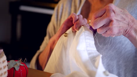 The-wrinkled-hands-of-an-aging-elderly-woman-sewing-a-button-onto-a-white-dress-shirt-with-thread