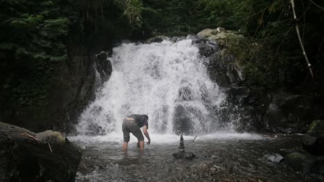Indonesian-man-building-cairns-balanced-rock-tower-in-a-river-in-front-of-a-waterfall