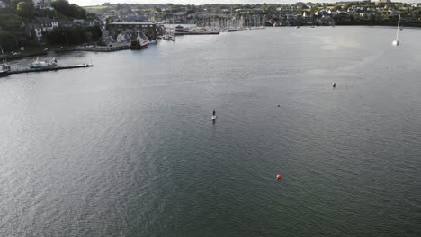 Drone-footage-of-a-SUP-boarder-paddling-on-a-wide-river,-passing-fishing-boats-and-town