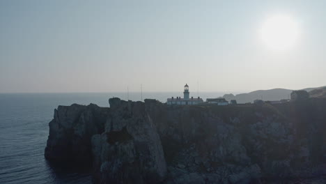 Slow-aerial-push-back-shot-revealing-Gamov-lighthouse-complex-standing-on-a-rocky-steep-cliff,-on-the-sunset