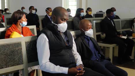 Small-church-of-people-social-distancing-and-watching-the-somber-funeral-service,-whilst-sitting-on-benches-and-wearing-protective-face-masks-against-Covid-19