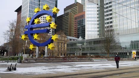 Adult-Man-Wearing-Facemask-During-Pandemic-In-Front-Of-Euro-Sign-Sculpture-In-Willy-Brandt-Platz-At-Wintertime-In-Frankfurt-am-Main,-Germany